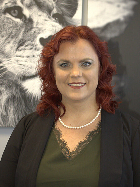 A woman with red hair wearing a black blazer and a green top, with a monochrome wildlife picture in the background.