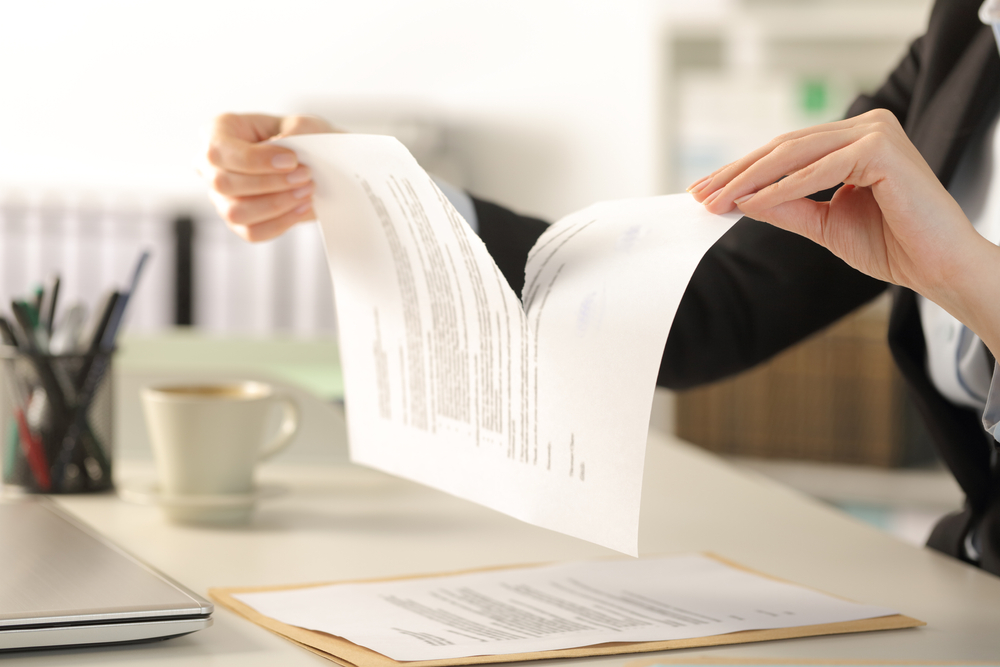 Person tearing a document in half at a desk with a cup of coffee, an envelope, and a laptop nearby, suggesting the cancellation or termination of a contract