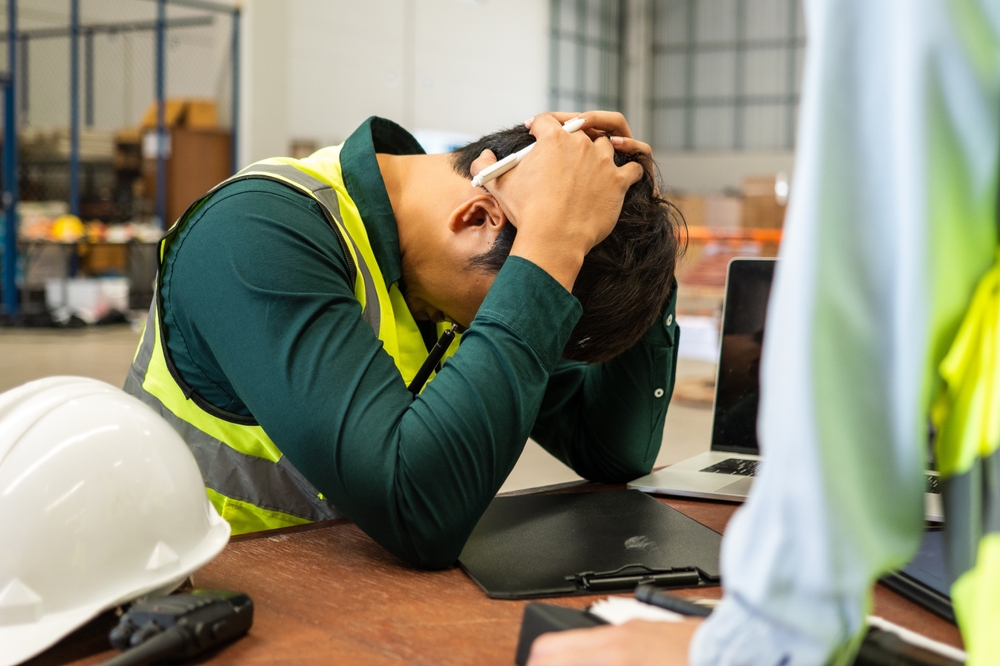 A construction worker wearing a safety vest sits at a desk, holding his head in frustration, with a hard hat and a laptop in front of him