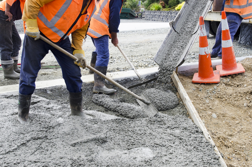 Construction workers in safety vests and boots spread wet concrete with shovels as it pours from a cement truck chute onto a prepared surface