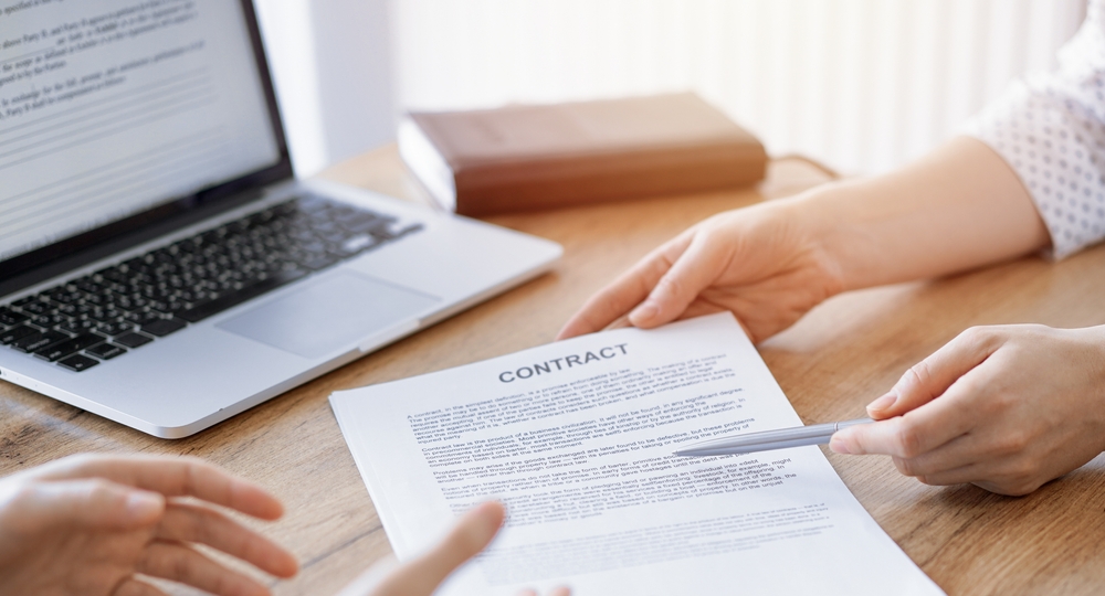 Two people discussing a contract document on a wooden table, with one person pointing to it using a pen. A laptop and book are visible in the background