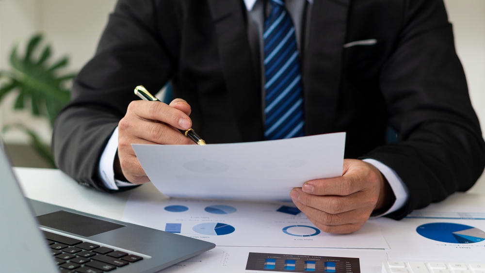 Business professional in a black suit and blue striped tie reviewing a document with charts at a desk, holding a pen. A laptop and financial reports are visible