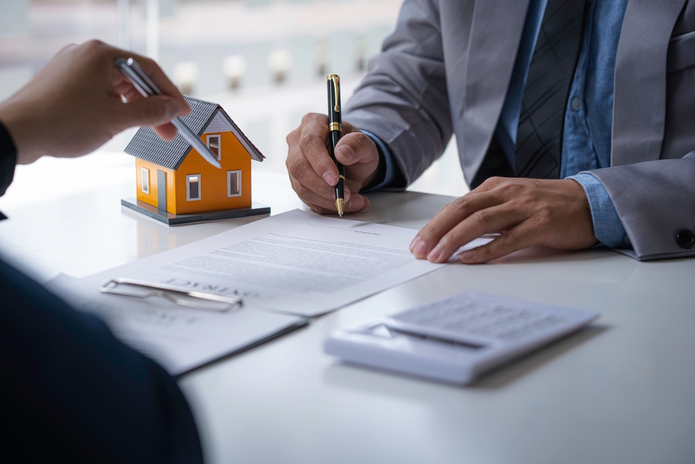 Person signing a real estate contract at a desk, with a small model house and a pen nearby, symbolizing property transactions or agreements