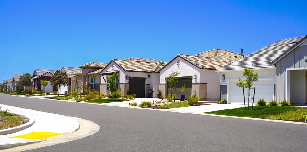 Suburban street with modern single-story homes, each with well-maintained lawns, driveways, and light-colored exteriors under a clear blue sky