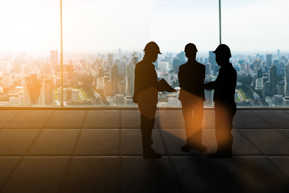 Three silhouetted construction workers discuss plans in front of a large window overlooking a city skyline at sunset
