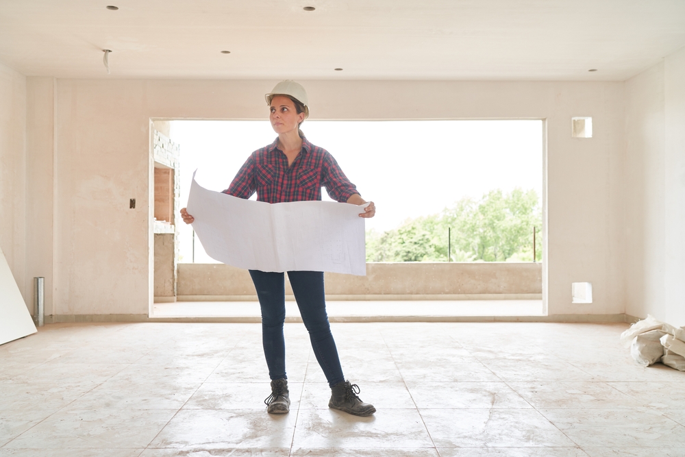 Woman in hard hat holding blueprints inside an unfinished room with large window and tiled floor