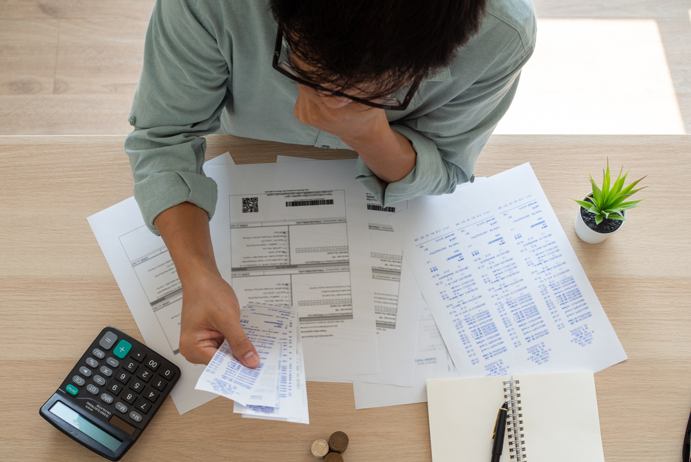 Person reviewing bills and receipts at a desk with papers, a calculator, coins, a notebook, pen, and a small potted plant