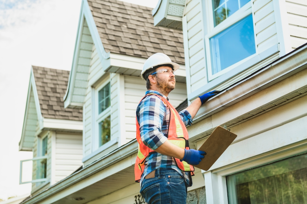 Construction worker in safety gear inspecting a house roof and gutter, holding a clipboard, likely performing a home inspection