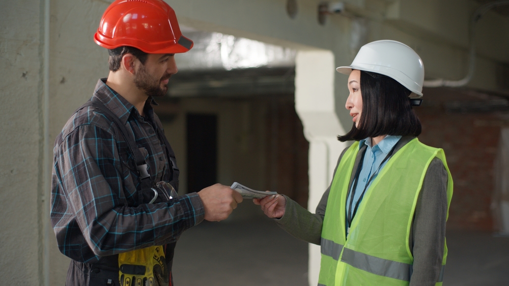 Man in a red hard hat hands documents to a woman in a white hard hat and safety vest inside a construction site