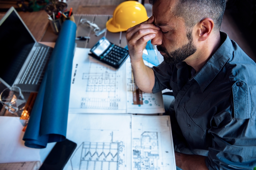 Stressed man at a desk with blueprints, laptop, and hard hat, pinching his nose bridge in frustration or fatigue